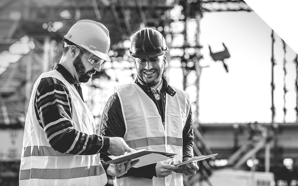 Zwei Bauarbeiter mit Helmen und Westen betrachten gemeinsam Dokumente für die Jährliche Unterweisung auf einer Baustelle.